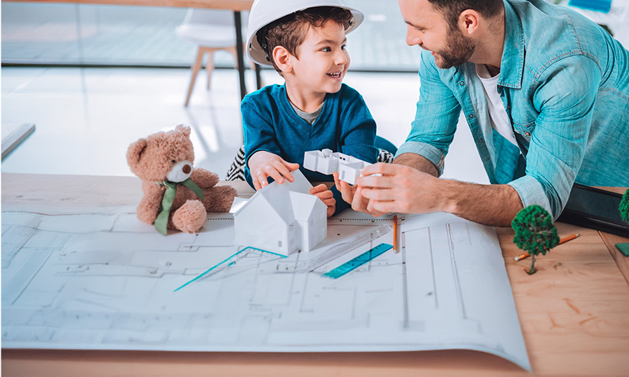 Auf dem Bild ist ein kleiner Junge zu sehen, der einen weißen Bauhelm trägt. Links neben dem Jungen befindet sich ein Teddybär. Rechts neben Ihm ein Junger Mann. Vor den beiden befindet sich ein aus Papier gebautes Haus. 