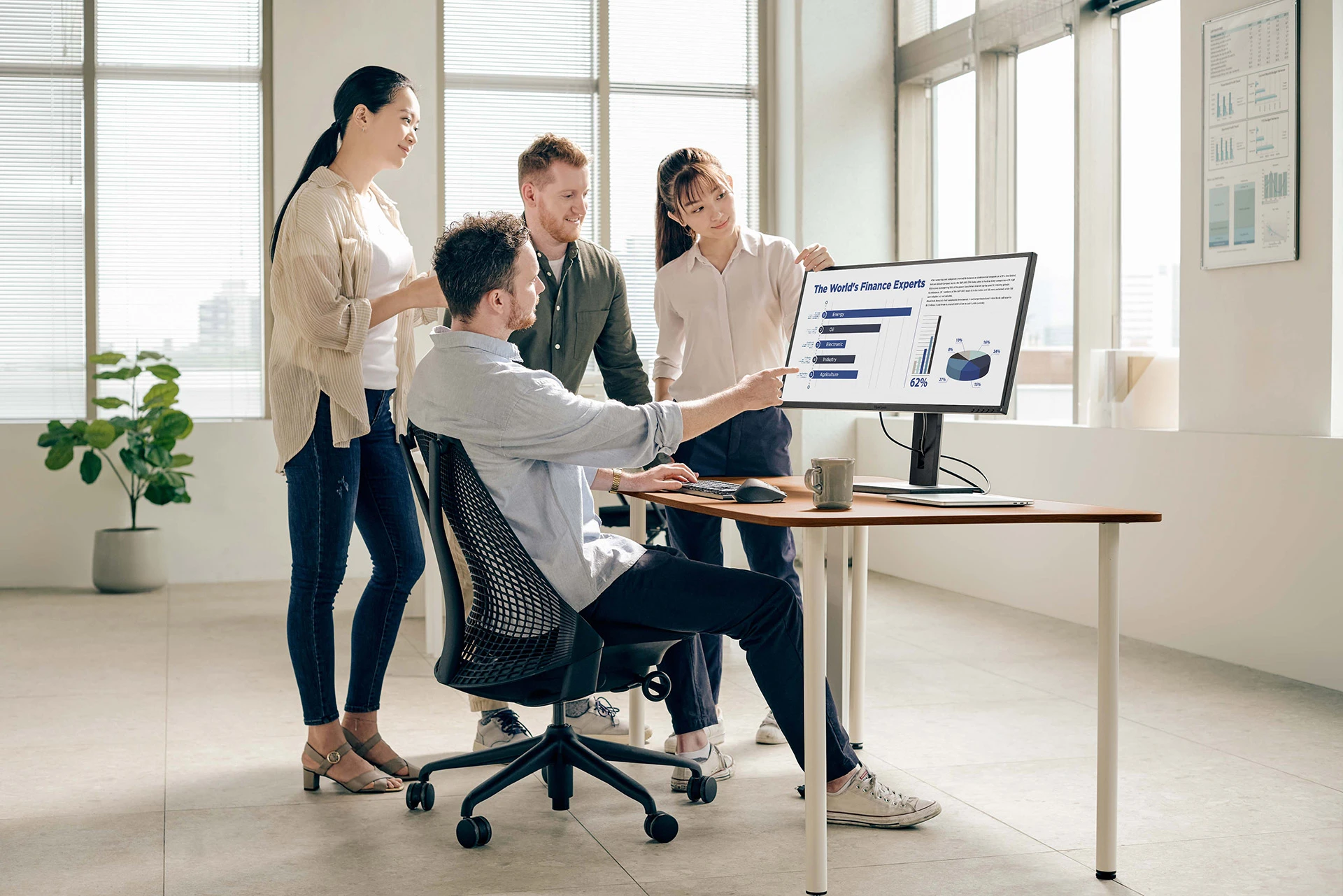 An einem Schreibtisch mit Monitor sitzt ein Mann, der auf eine Statistik zeigt, welche auf dem Monitor angezeigt wird. Um ihn herum stehen drei weitere Menschen, die sich die Statistik anschauen und sich über die Zahlen austauschen.