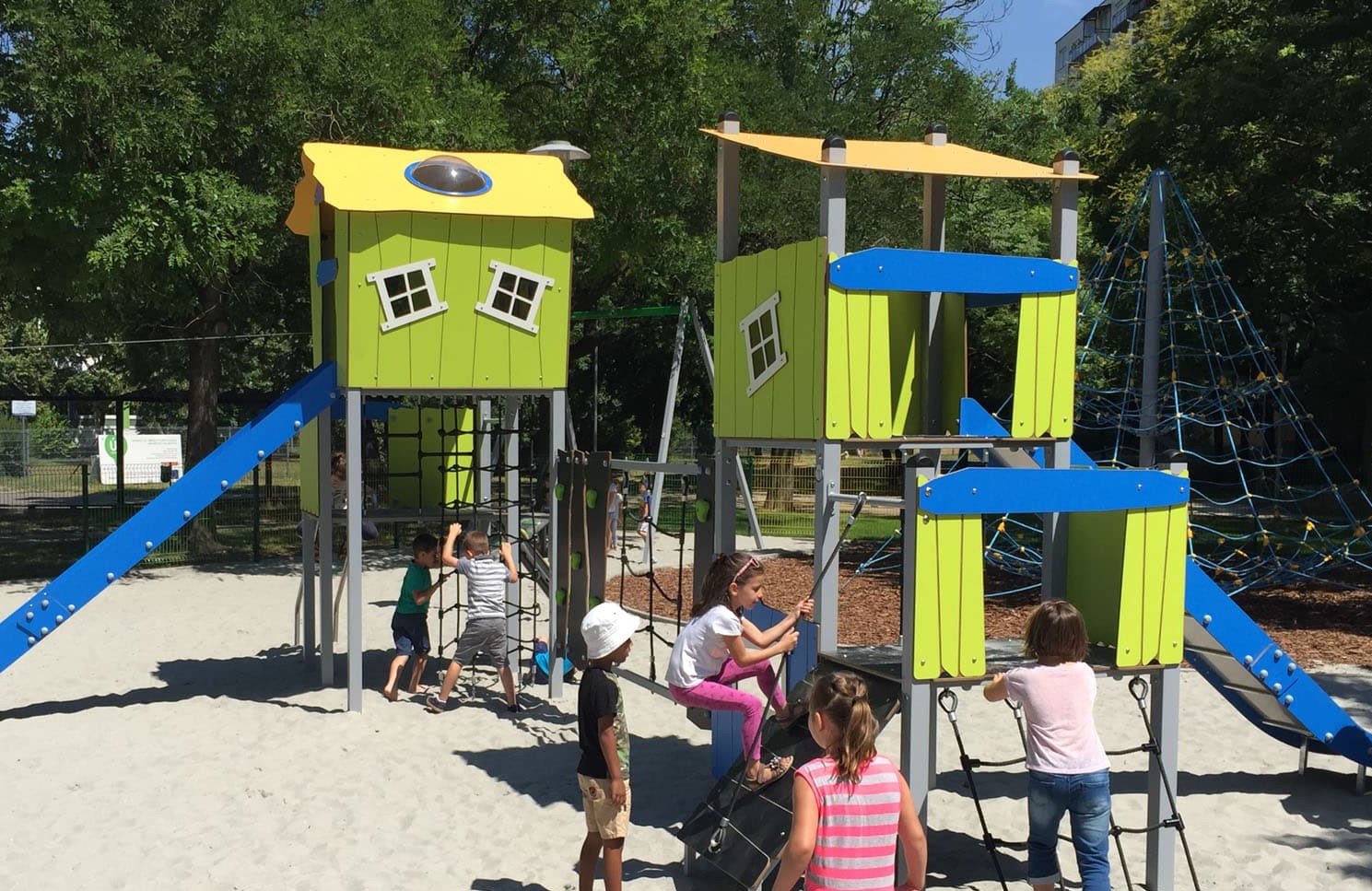 Ein grün-blaues Spielhaus mit zwei großen Dächern und zwei Rutschen steht auf einem Sandplatz. Um das Spielgerüst stehen viele Kinder.