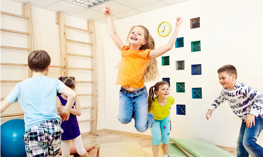 In einem Raum spielen fünf Kinder. Ein Mädchen springt in der Mitte in die Luft und wirft die Arme nach oben.