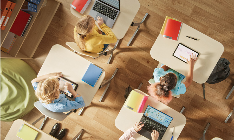 Das Bild zeigt einen Klassenraum von oben. Zu sehen sind vier Tische und Schüler, die an Ihren digitalen Endgeräten arbeiten.