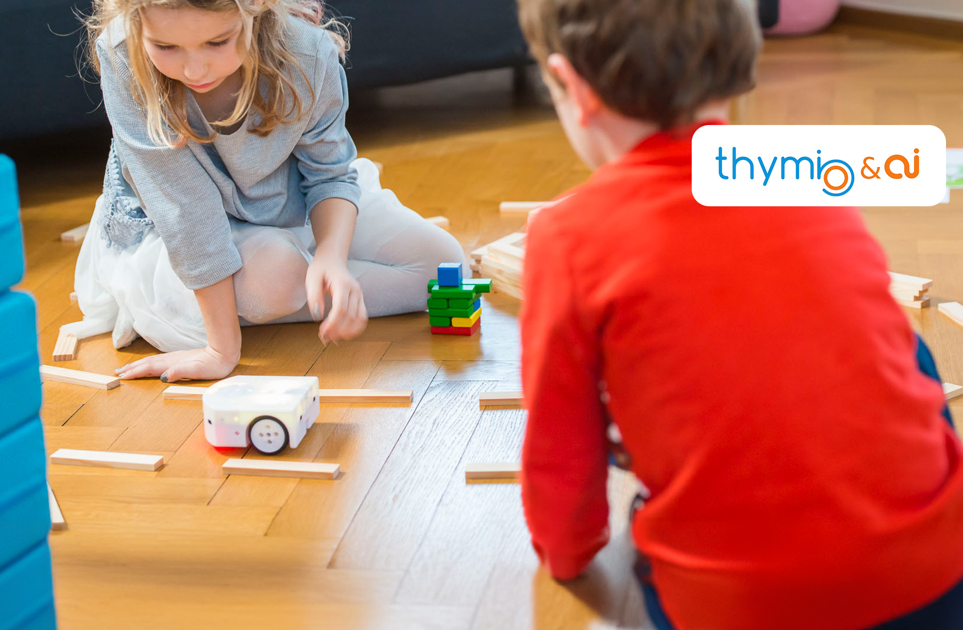 Zwei Kinder spielen mit dem Thymio Lernroboter auf dem Boden und lassen diesen durch einen Parcours fahren. Oben rechts ist das Thymio Logo zu sehen.