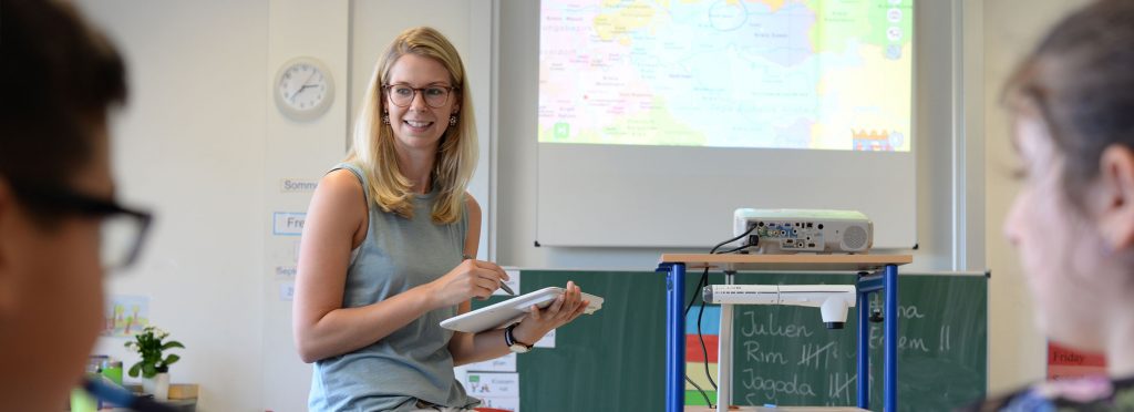 Lehrerin steht vorne mit einem Tablet in der Hand. Rechts neben ihre befindet eine Dokumentenkamera, sowie ein Beamer, dessen Bild an die Wand projiziert wird. 