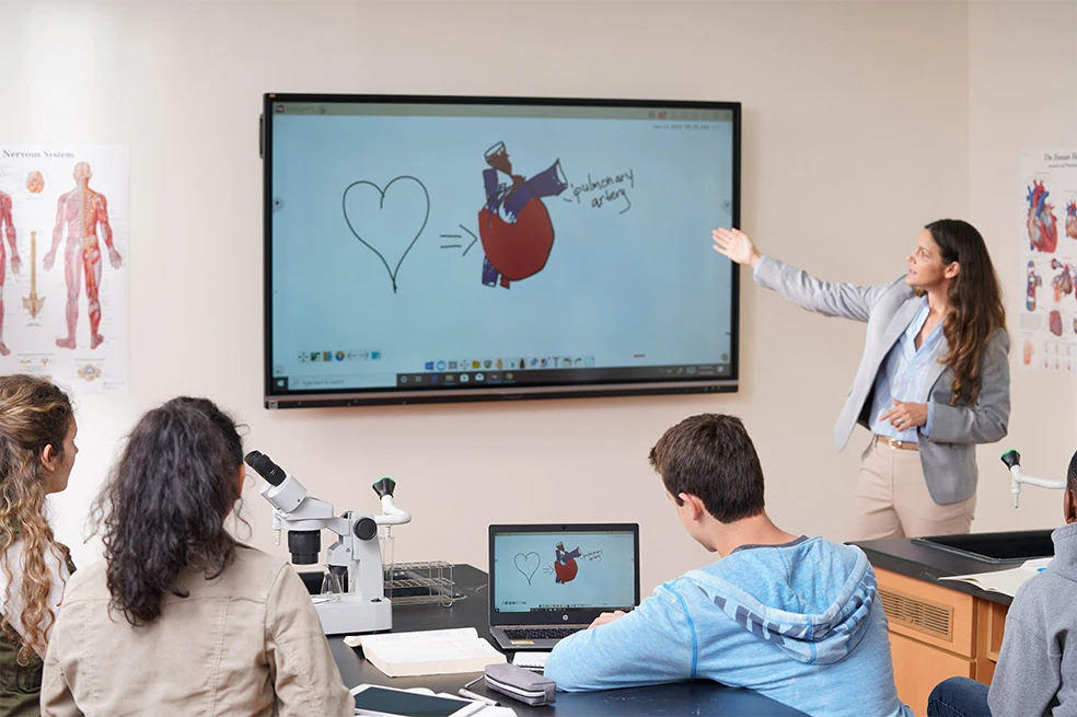 In einem Klassenraum steht eine Lehrerin vorne an einem Display von ViewSonic und erklärt eine Aufgabe. Die Schüler im Klassenraum hören der Lehrerin dabei zu