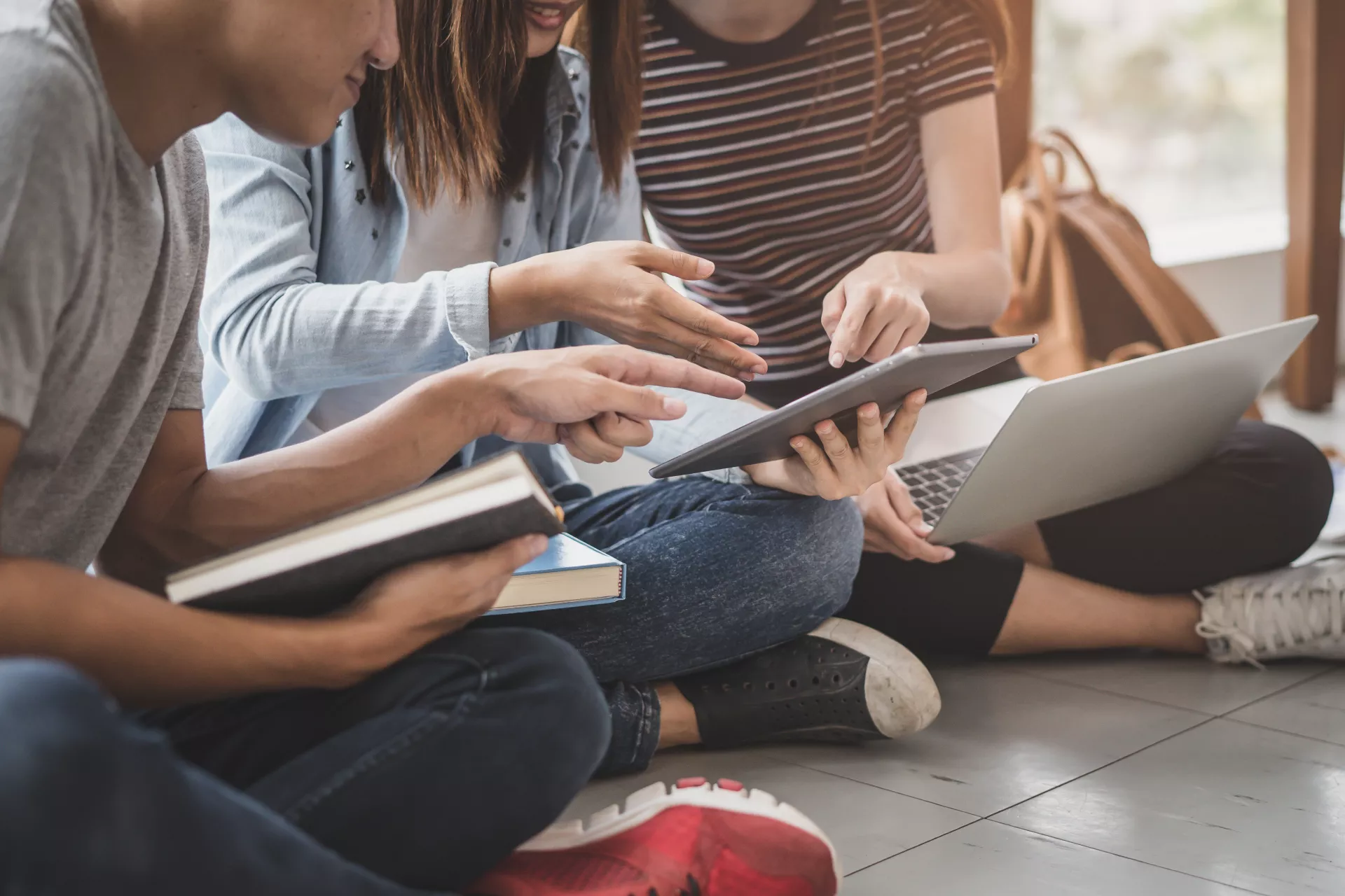 Drei Personen sitzen auf dem Boden und halten einen Laptop, ein Tablet und ein Buch in der Hand