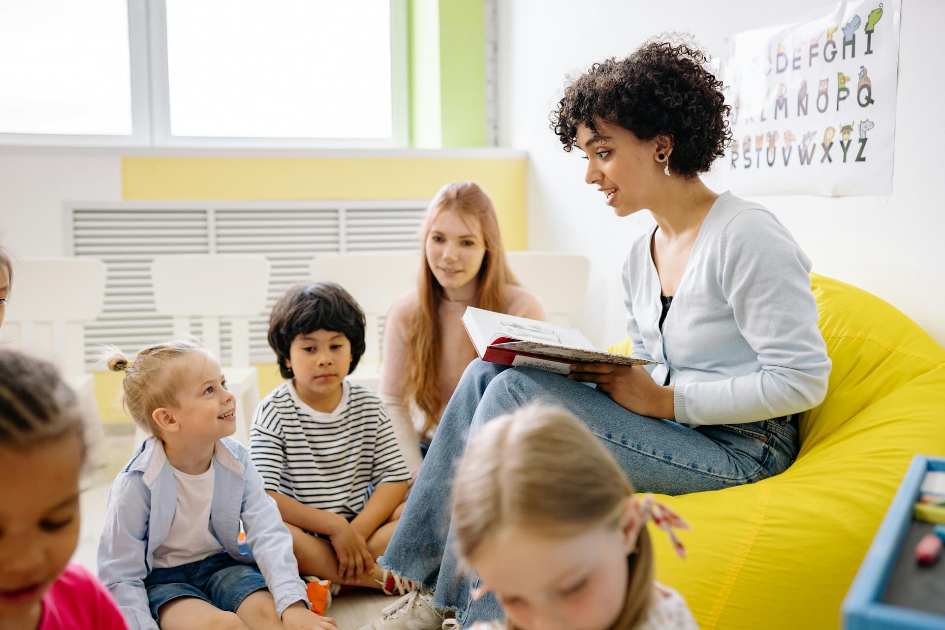 Frau sitzt auf einem gelben Sitzsack und liest Kindern aus einem Buch vor