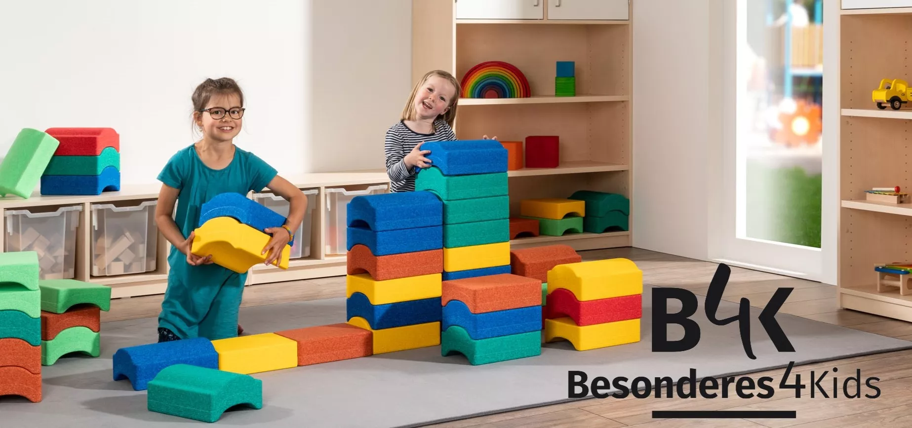 Zwei Mädchen spielen mit mehreren bunten Bausteinen. In der Ecke des Bildes ist ein Logo von Besonderes4Kids