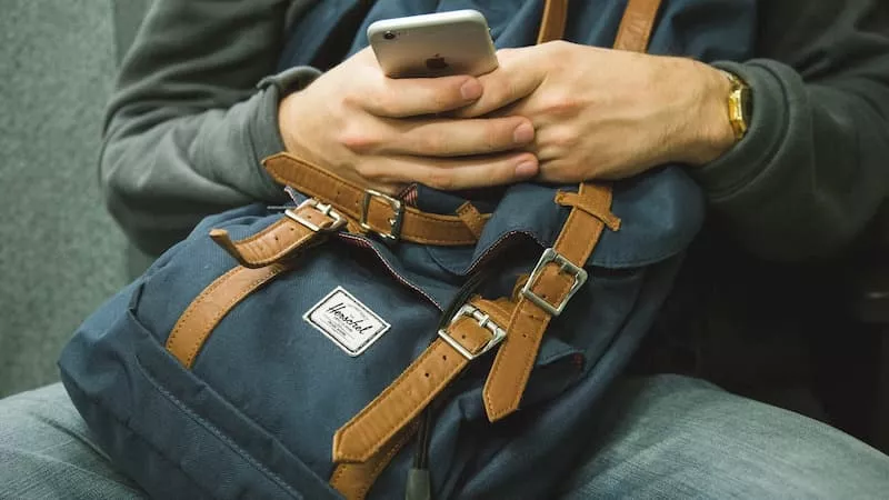 Person, die sitzt, mit einem blauen Rucksack auf dem Schoß und dem Handy in den Händen