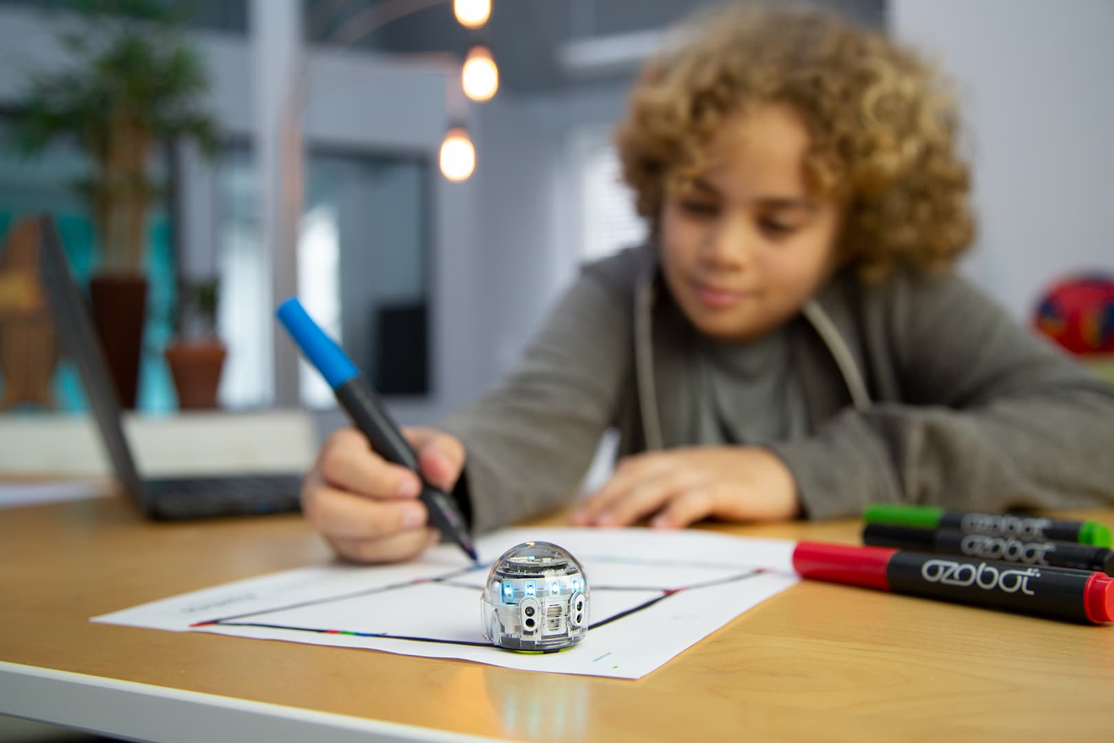 Ein Junge sitzt an einem Tisch und malt mit den Stiften von Ozobot Linien auf ein Blatt Papier. Im Vordergrund des Bildes steht ein kleiner Roboter von Ozobot auf dem Blatt Papier.