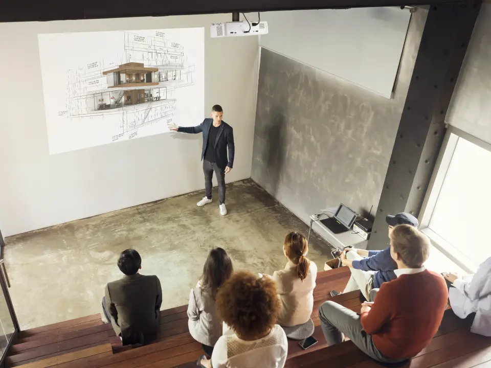 In einem Hörsaal sitzen ein paar Menschen, welche Ihren Blick nach vorne auf eine Leinwand gerichtet haben. Vor dieser Leinwand steht ein Mann, der die Inhalte zeigt und erklärt. Die Inhalte werden von einem Beamer von ViewSonic auf die Leinwand projizier