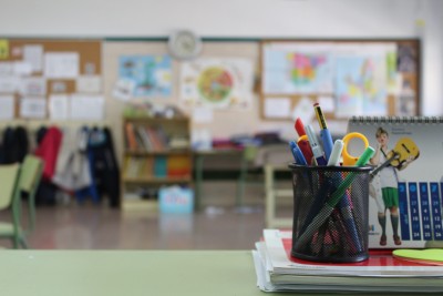 Nahaufnahme eines Schreibtisches. Fokus auf einem Stiftebehälter, der auf Blöcken steht. Im Hintergrund ein Klassenzimmer