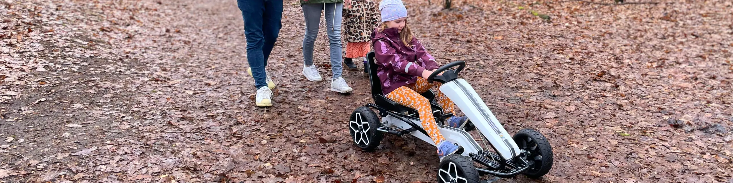 Mädchen sitzt auf einem Kettcar im Wald. Hinter ihr sind die Beine weiterer Personen zu sehen.