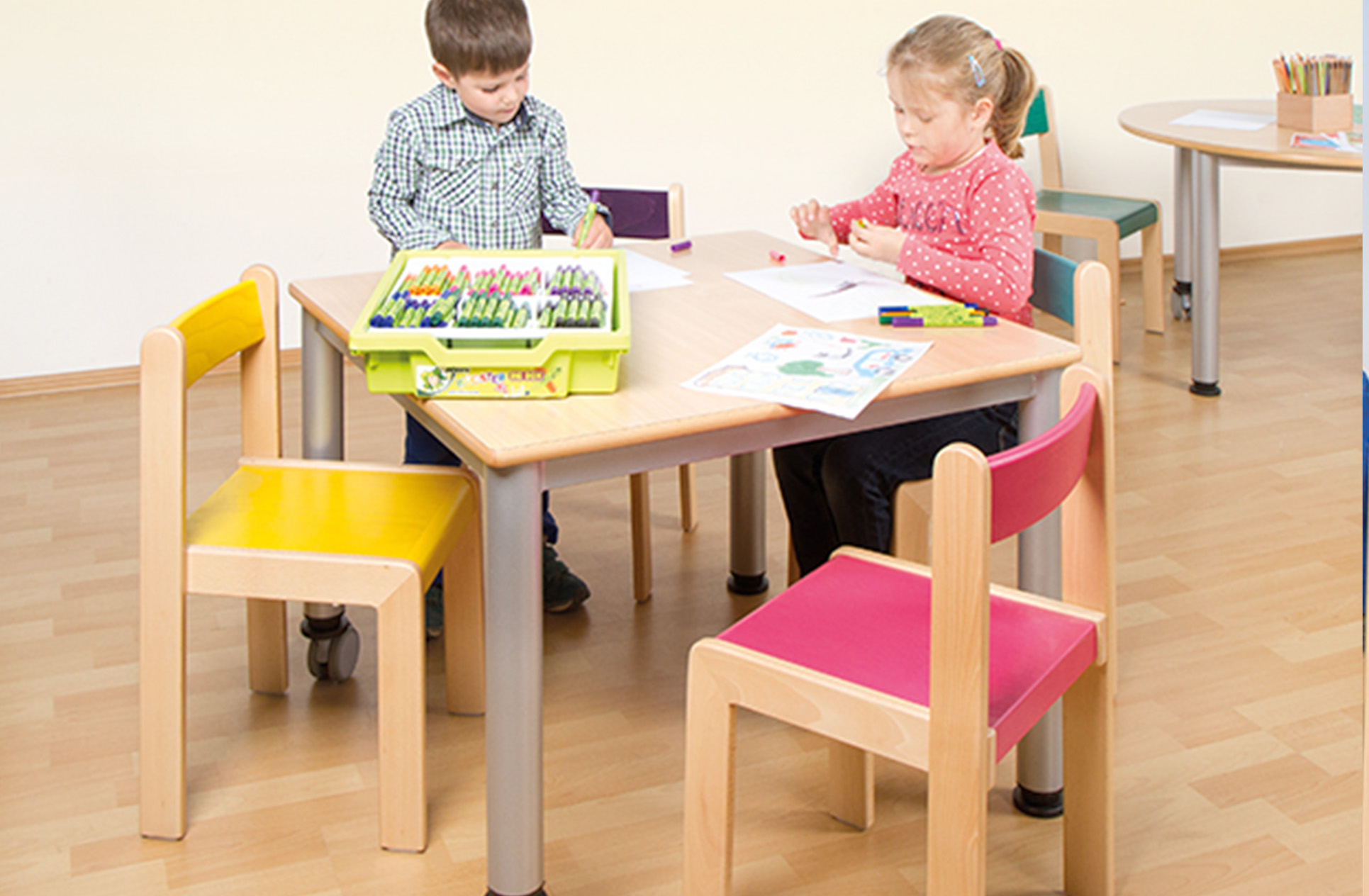 Zwei Kinder sitzen an einem quadratischen Tisch, auf zwei Stühlen mit bunter Sitzfläche. Auf dem Tisch liegen Stifte, sowie Blätter zum malen.