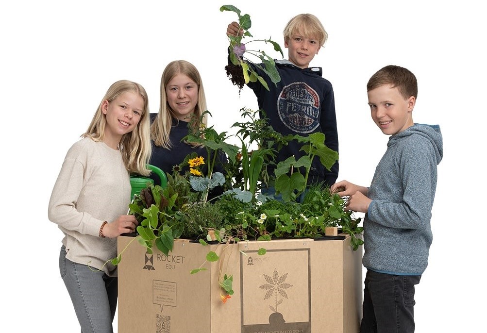 Vier Schüler stehen um das Rocket EDU In- und Outdoor Hochbeet "Perma-Kit" während einer der Schüler eine Pflanze aus dem Hochbeet hochhebt.