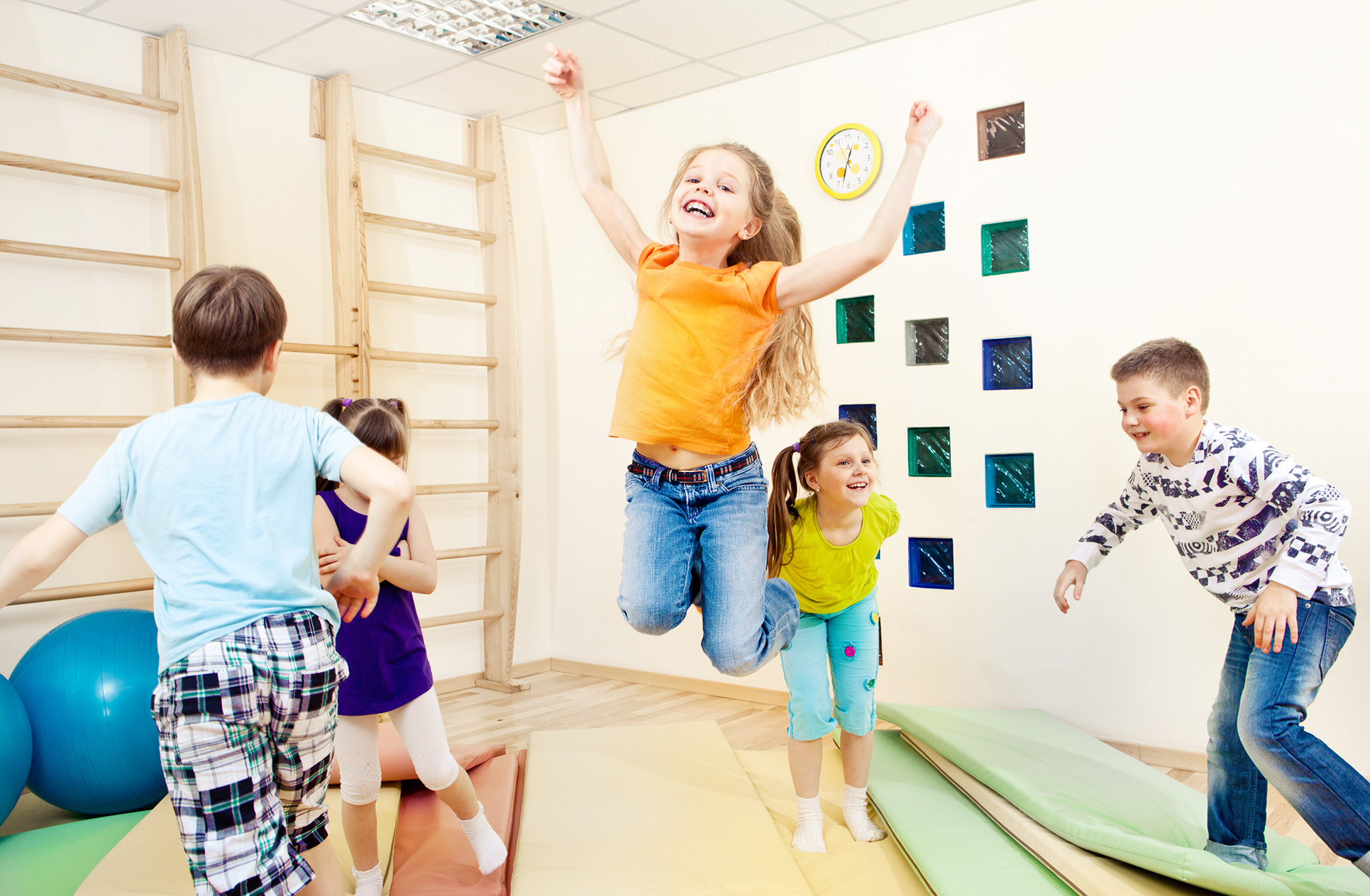 In einem Raum spielen fünf Kinder. Ein Mädchen springt in der Mitte in die Luft und wirft die Arme nach oben.